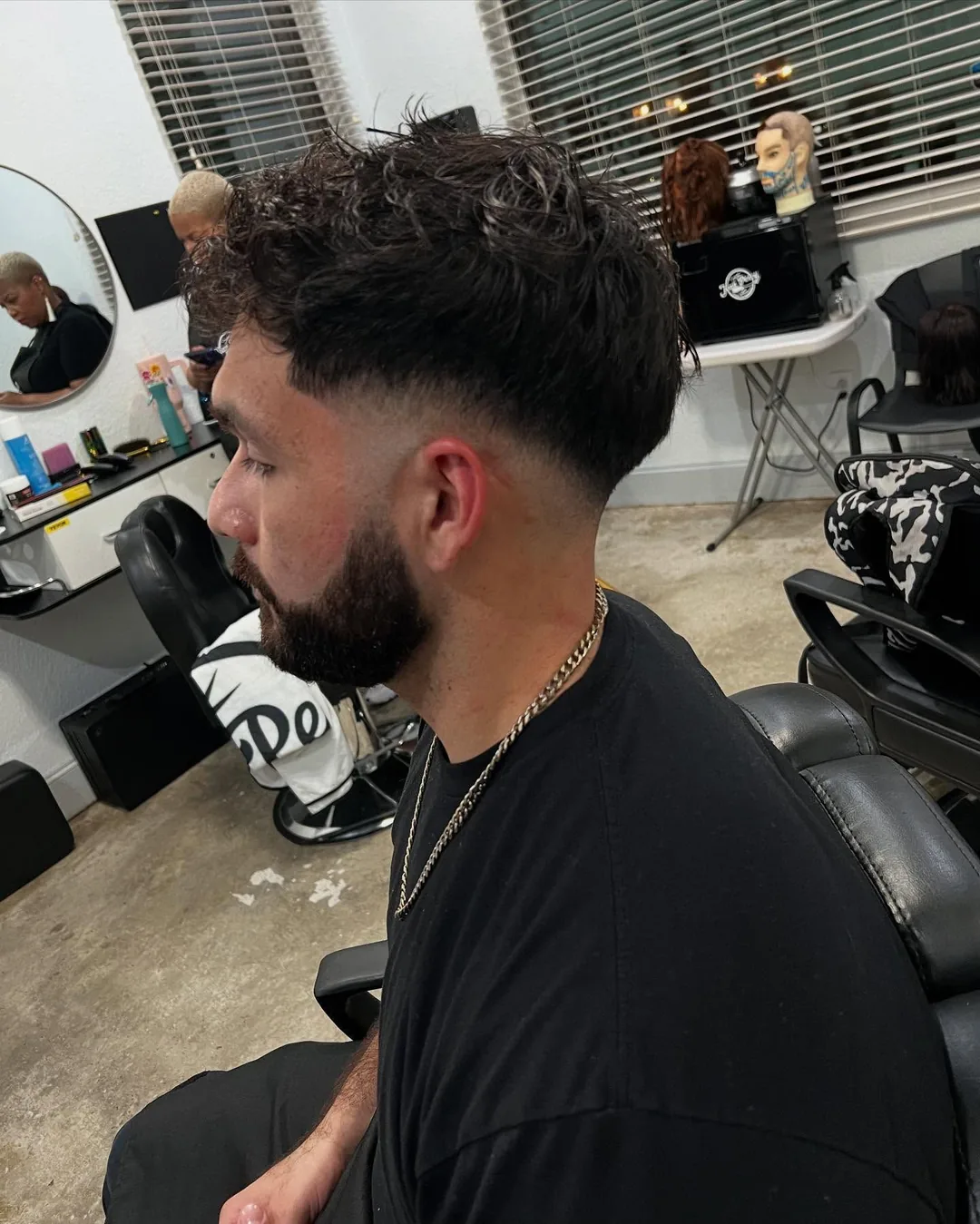 Profile of a man with dark curly hair and beard, seated in a barber chair in a salon.