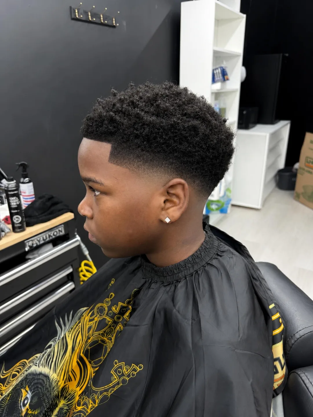 Boy with a short curly fade haircut in a barbershop, wearing a black cape with gold design.